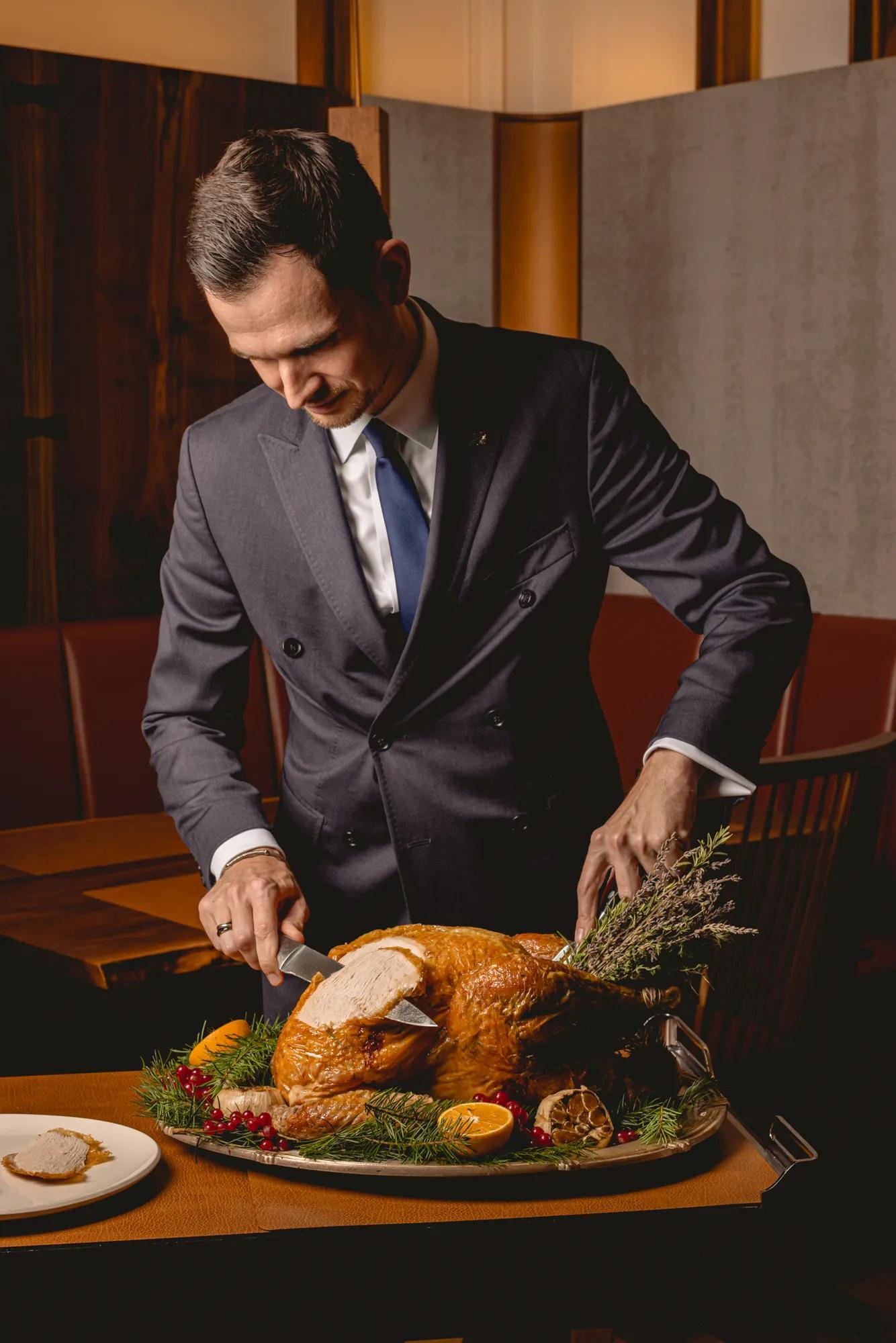 Server in a dark suit carving a golden roasted turkey garnished with herbs, citrus, and cranberries on a platter.