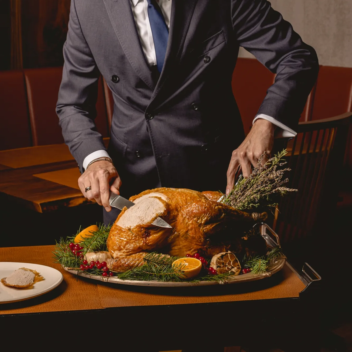 Server in a dark suit carving a golden roasted turkey garnished with herbs, citrus, and cranberries on a platter.