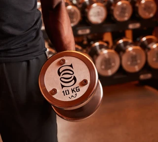 Close-up of a person holding a dumbbell with a 10 kg weight, displaying the Surrenne logo on the side of the dumbbell, with other dumbbells in the background.