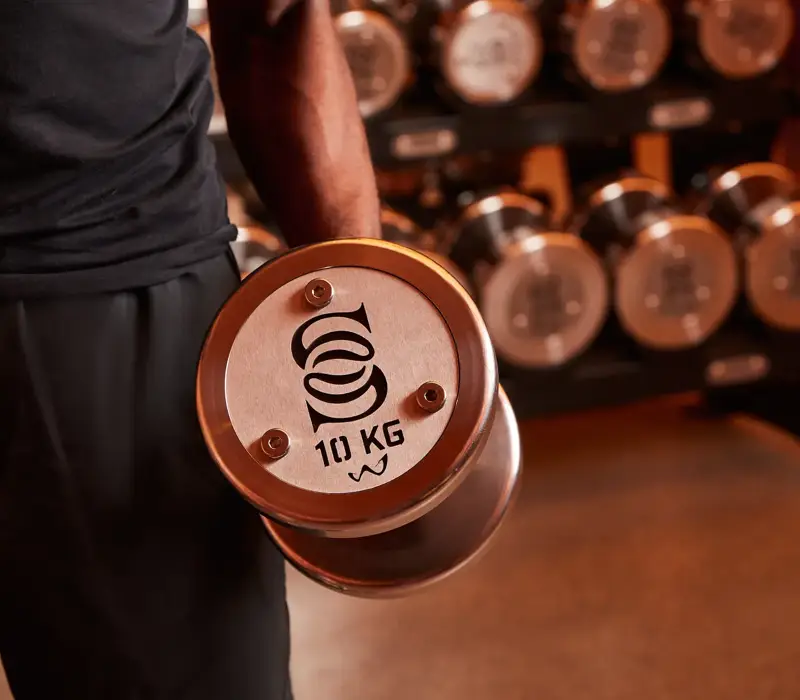Close-up of a person holding a dumbbell with a 10 kg weight, displaying the Surrenne logo on the side of the dumbbell, with other dumbbells in the background.