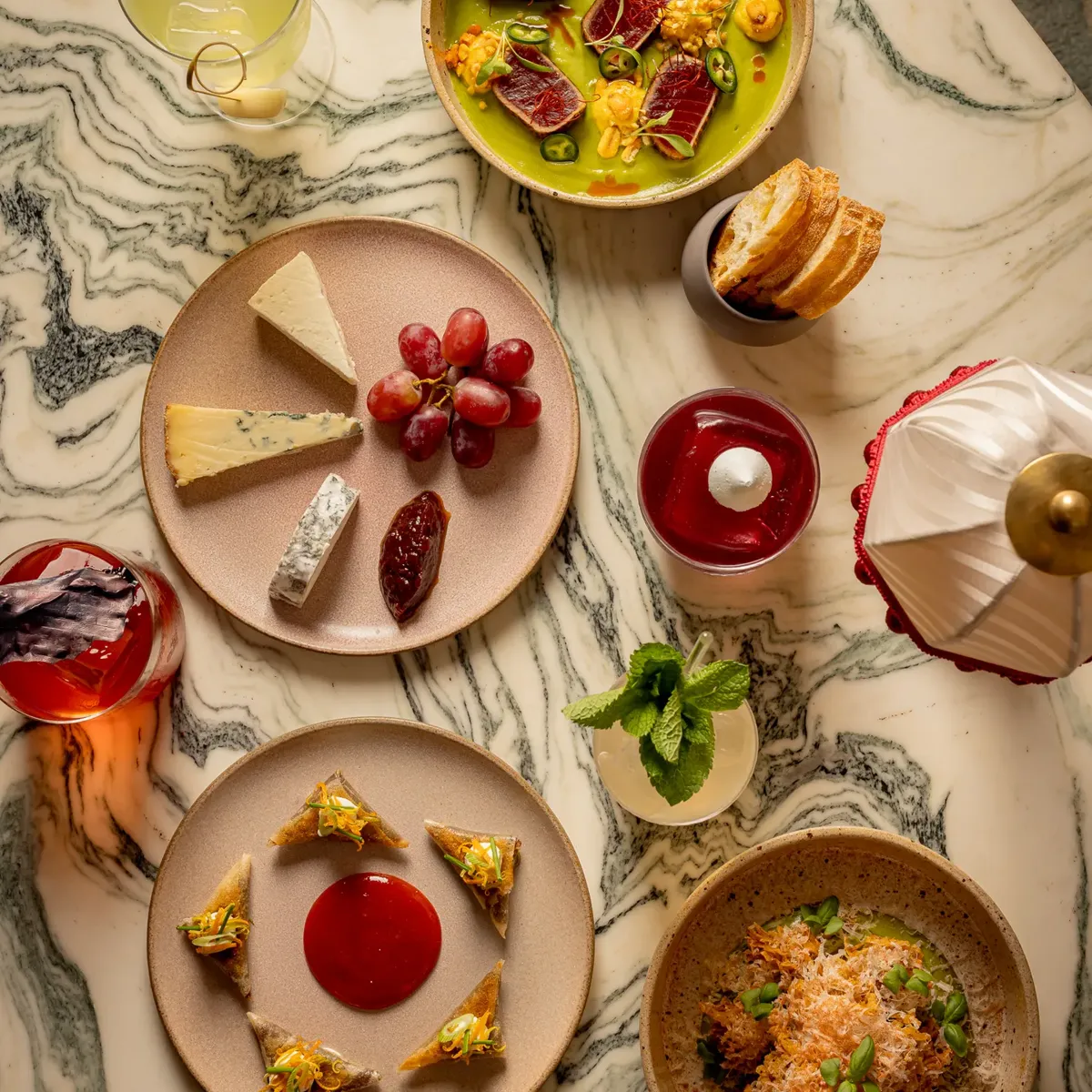 Selection of colourful small plates and drinks arranged on a marbled table, including cheese, grapes, soup, and vibrant desserts.