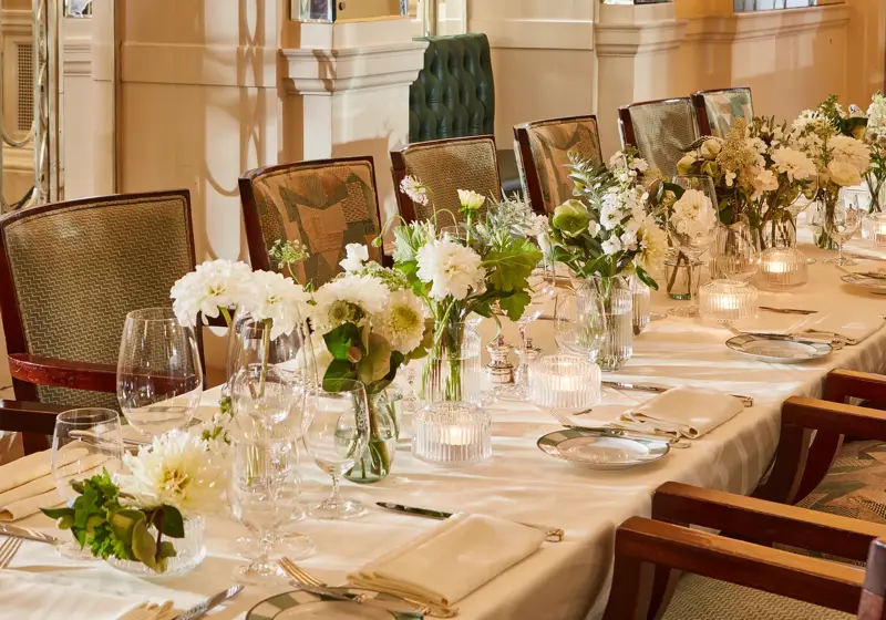 Elegant banquet table set with white floral arrangements, glassware, candles, and upholstered chairs in a refined dining room.