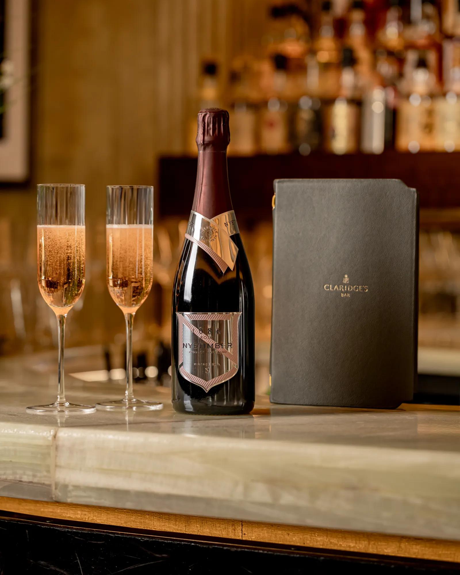 Champagne bottle, two filled flutes and a Claridge’s Bar menu displayed on a marble bar counter with softly lit shelves in the background.