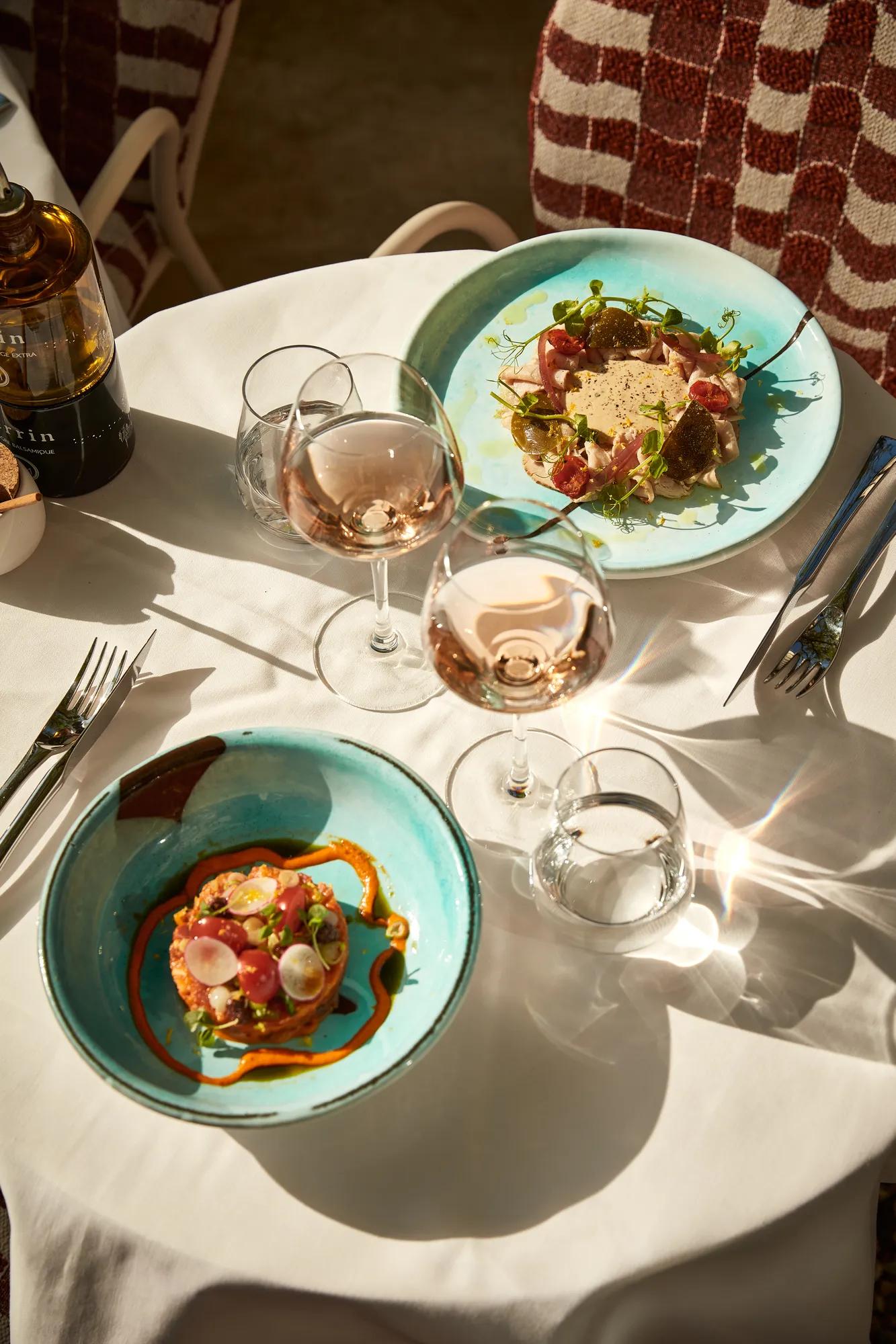 Two turquoise plates of gourmet dishes with rosé wine glasses on a sunlit terrace table.