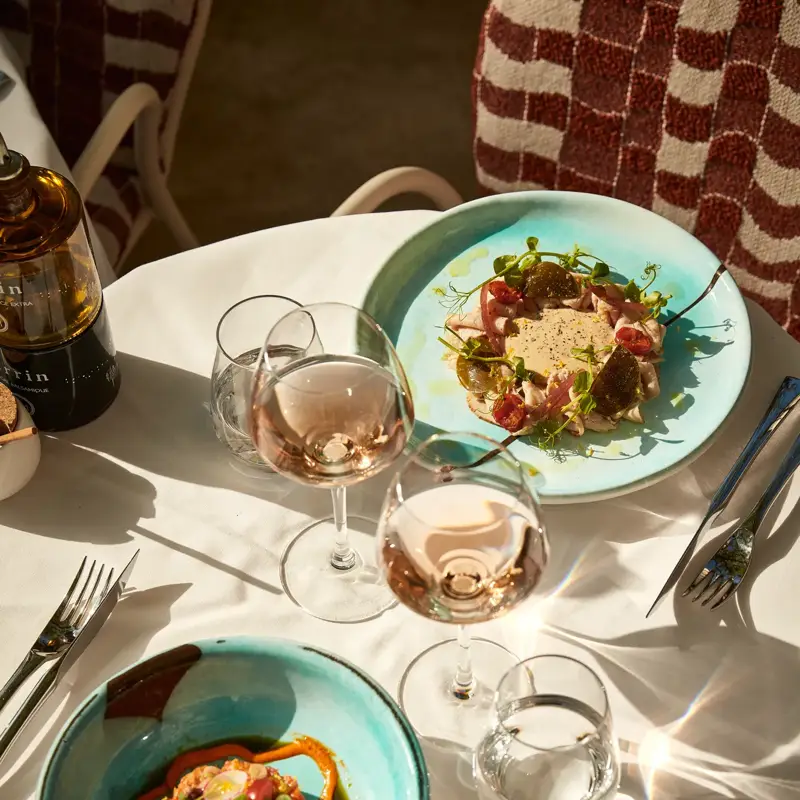 Two turquoise plates of gourmet dishes with rosé wine glasses on a sunlit terrace table.