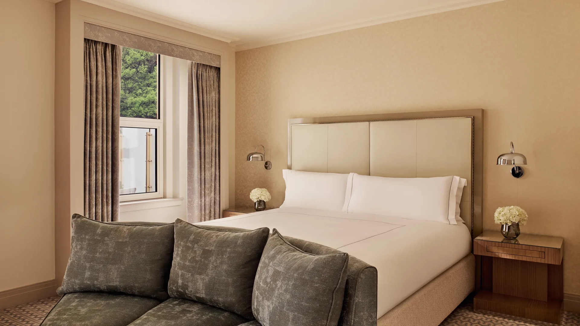 Neutral bedroom with king bed, green sofa, and window view framed by beige curtains.