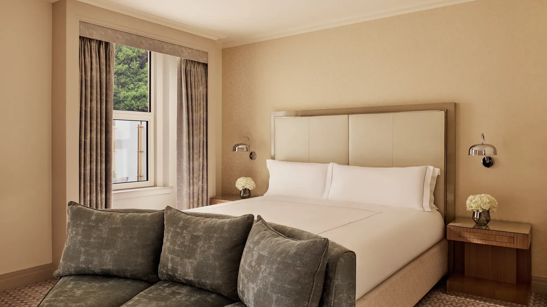 Neutral bedroom with king bed, green sofa, and window view framed by beige curtains.