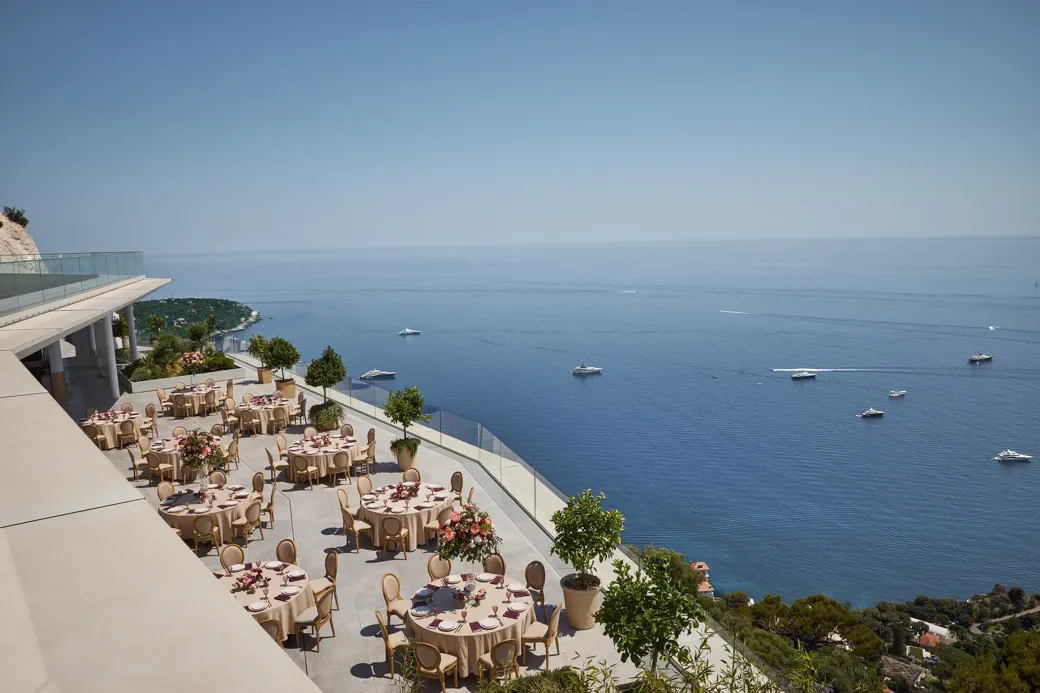 Terrasse extérieure élégante aménagée pour un événement, avec des tables rondes couvertes de nappes beiges et décorées de centres de table floraux roses. La terrasse offre une vue imprenable sur la mer, parsemée de yachts. Des garde-corps en verre et des arbres en pot bordent le rebord, et le littoral est visible au loin sous un ciel bleu clair.