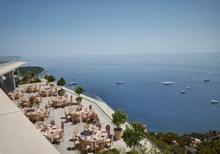 Terrasse extérieure élégante aménagée pour un événement, avec des tables rondes couvertes de nappes beiges et décorées de centres de table floraux roses. La terrasse offre une vue imprenable sur la mer, parsemée de yachts. Des garde-corps en verre et des arbres en pot bordent le rebord, et le littoral est visible au loin sous un ciel bleu clair.