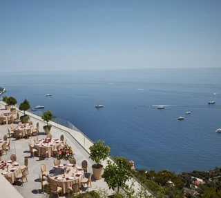 Terrasse extérieure élégante aménagée pour un événement, avec des tables rondes couvertes de nappes beiges et décorées de centres de table floraux roses. La terrasse offre une vue imprenable sur la mer, parsemée de yachts. Des garde-corps en verre et des arbres en pot bordent le rebord, et le littoral est visible au loin sous un ciel bleu clair.