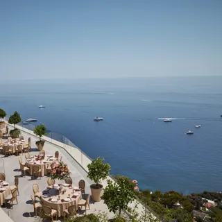 Terrasse extérieure élégante aménagée pour un événement, avec des tables rondes couvertes de nappes beiges et décorées de centres de table floraux roses. La terrasse offre une vue imprenable sur la mer, parsemée de yachts. Des garde-corps en verre et des arbres en pot bordent le rebord, et le littoral est visible au loin sous un ciel bleu clair.