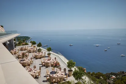 Terrasse extérieure élégante aménagée pour un événement, avec des tables rondes couvertes de nappes beiges et décorées de centres de table floraux roses. La terrasse offre une vue imprenable sur la mer, parsemée de yachts. Des garde-corps en verre et des arbres en pot bordent le rebord, et le littoral est visible au loin sous un ciel bleu clair.