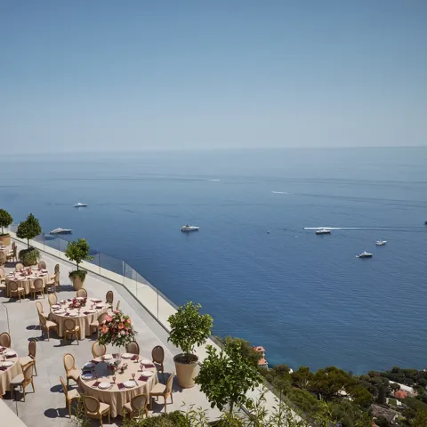 Terrasse extérieure élégante aménagée pour un événement, avec des tables rondes couvertes de nappes beiges et décorées de centres de table floraux roses. La terrasse offre une vue imprenable sur la mer, parsemée de yachts. Des garde-corps en verre et des arbres en pot bordent le rebord, et le littoral est visible au loin sous un ciel bleu clair.