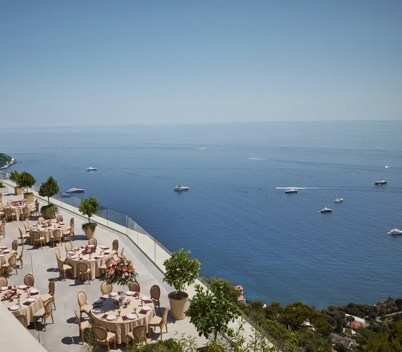 Elegant outdoor terrace set up for an event with round tables covered in beige tablecloths and decorated with pink floral centerpieces. The terrace overlooks a stunning view of the sea with several yachts scattered across the water. Glass railings and potted trees line the edge, and the coastline is visible in the distance under a clear blue sky.