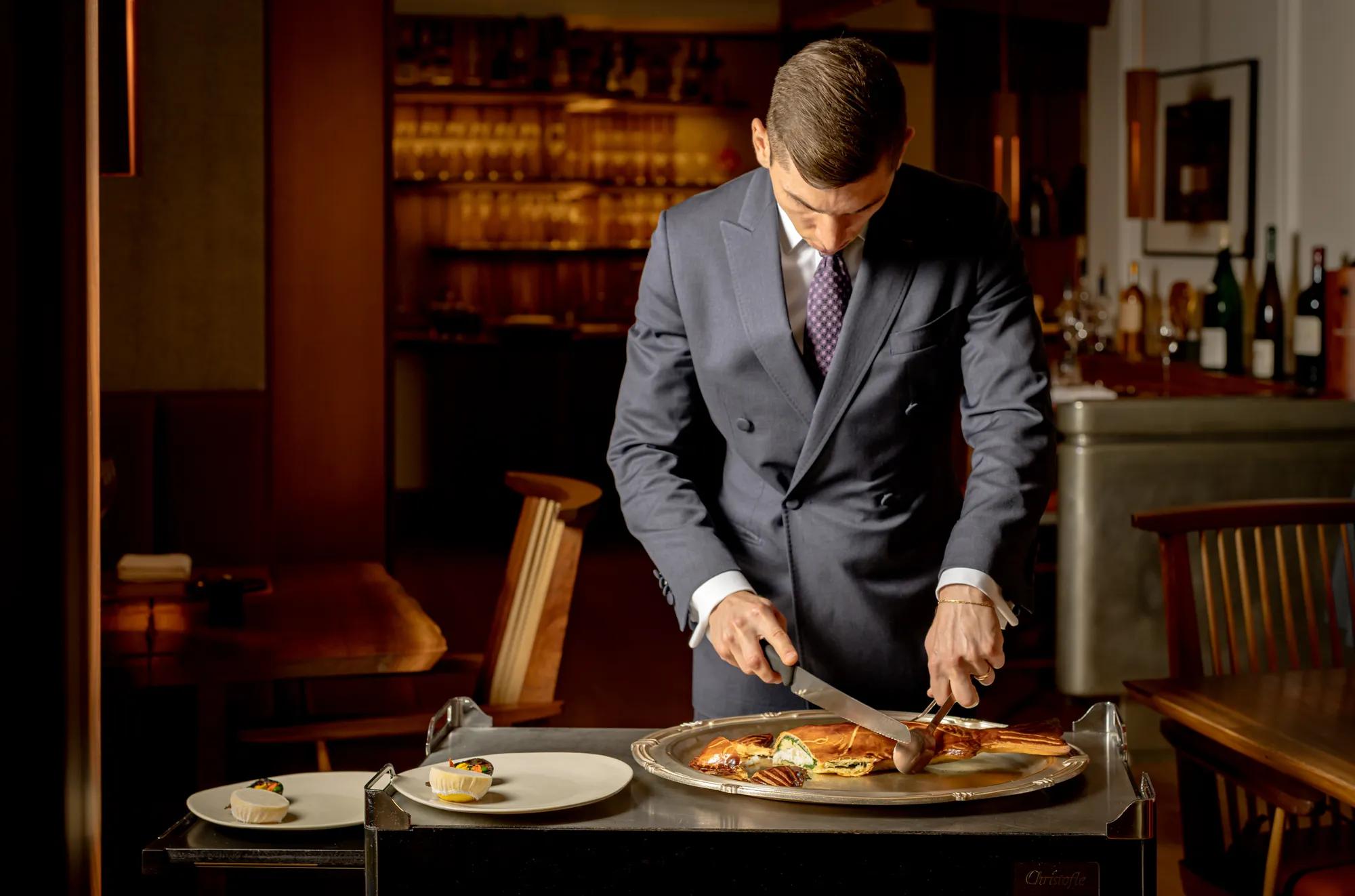 Person carving a cooked fish at a serving trolley in a restaurant dining room.
