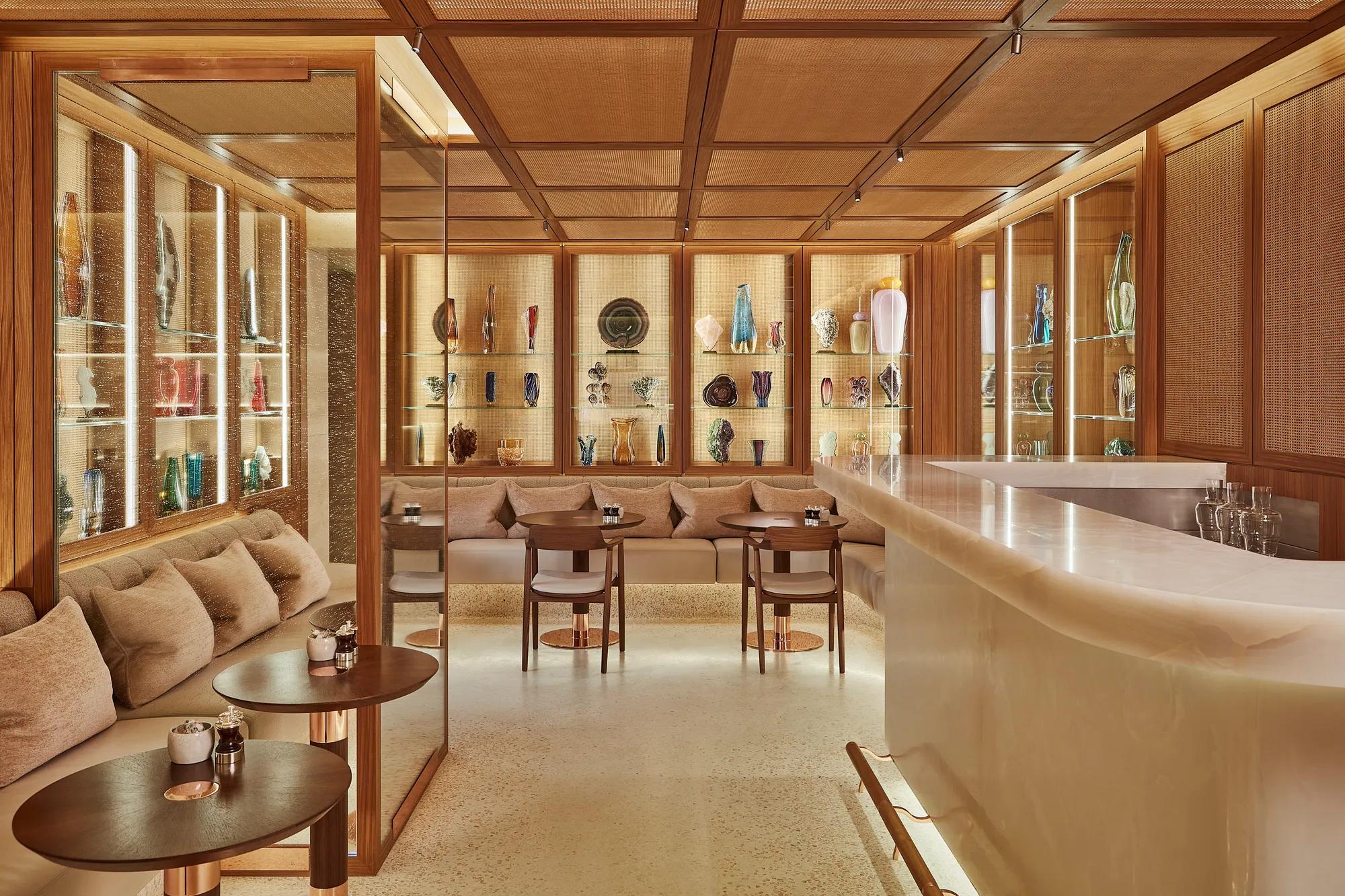 Elegant lounge bar with cushioned seating, marble counter, and illuminated glass shelves displaying colourful decorative vases and art pieces in a warm wood setting.