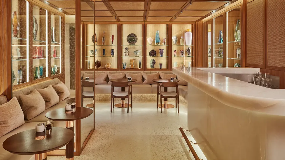 Elegant lounge bar with cushioned seating, marble counter, and illuminated glass shelves displaying colourful decorative vases and art pieces in a warm wood setting.