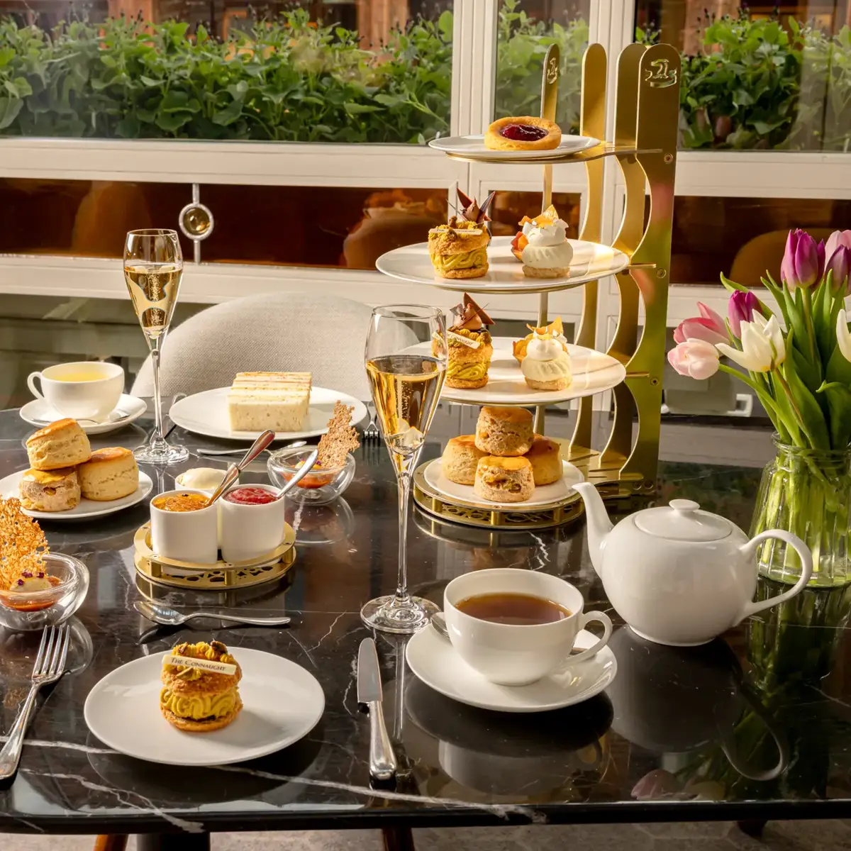 Afternoon tea spread with sandwiches, scones, pastries, tea, and champagne on a marble table beside tulips.