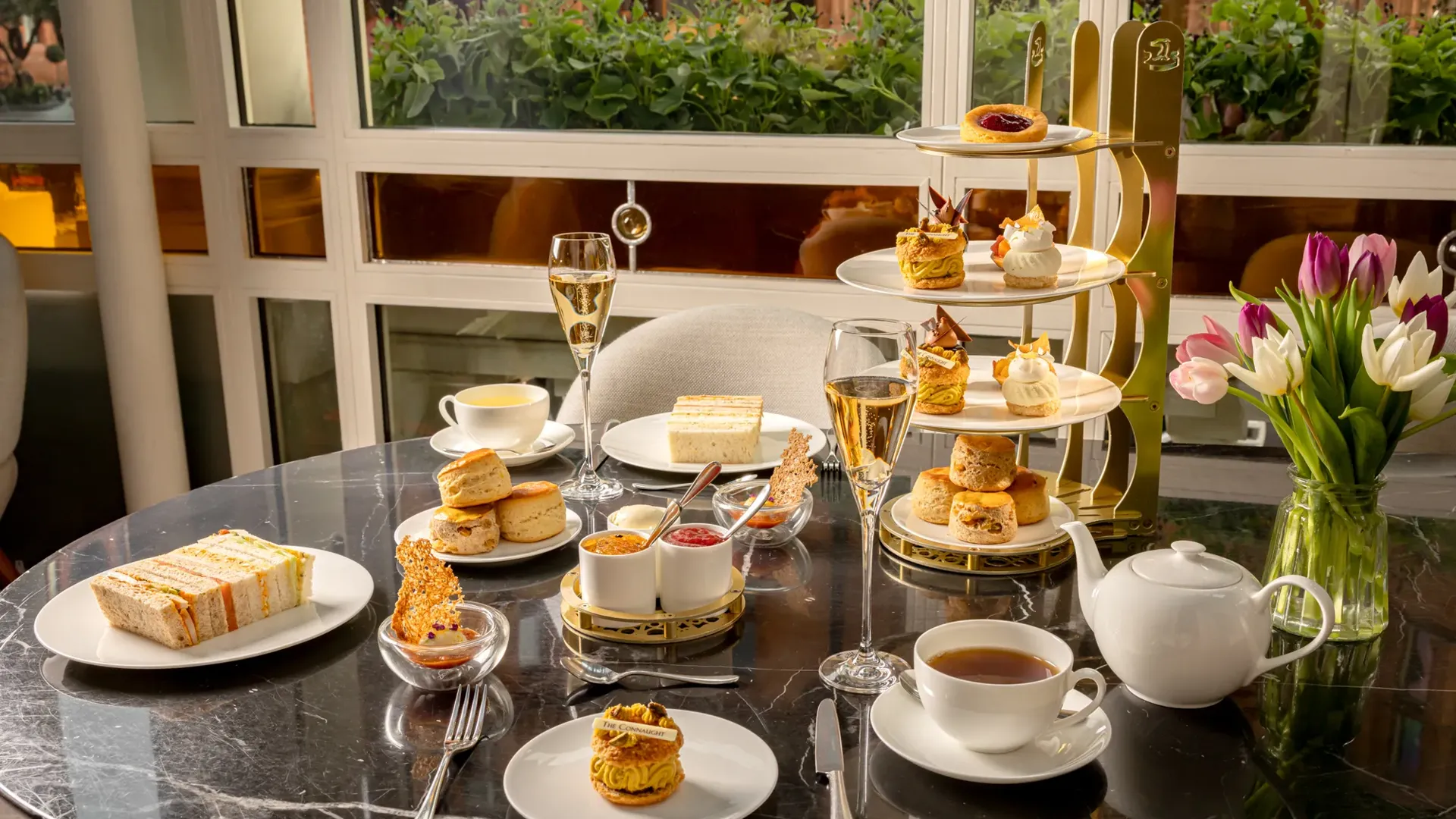 Afternoon tea spread with sandwiches, scones, pastries, tea, and champagne on a marble table beside tulips.