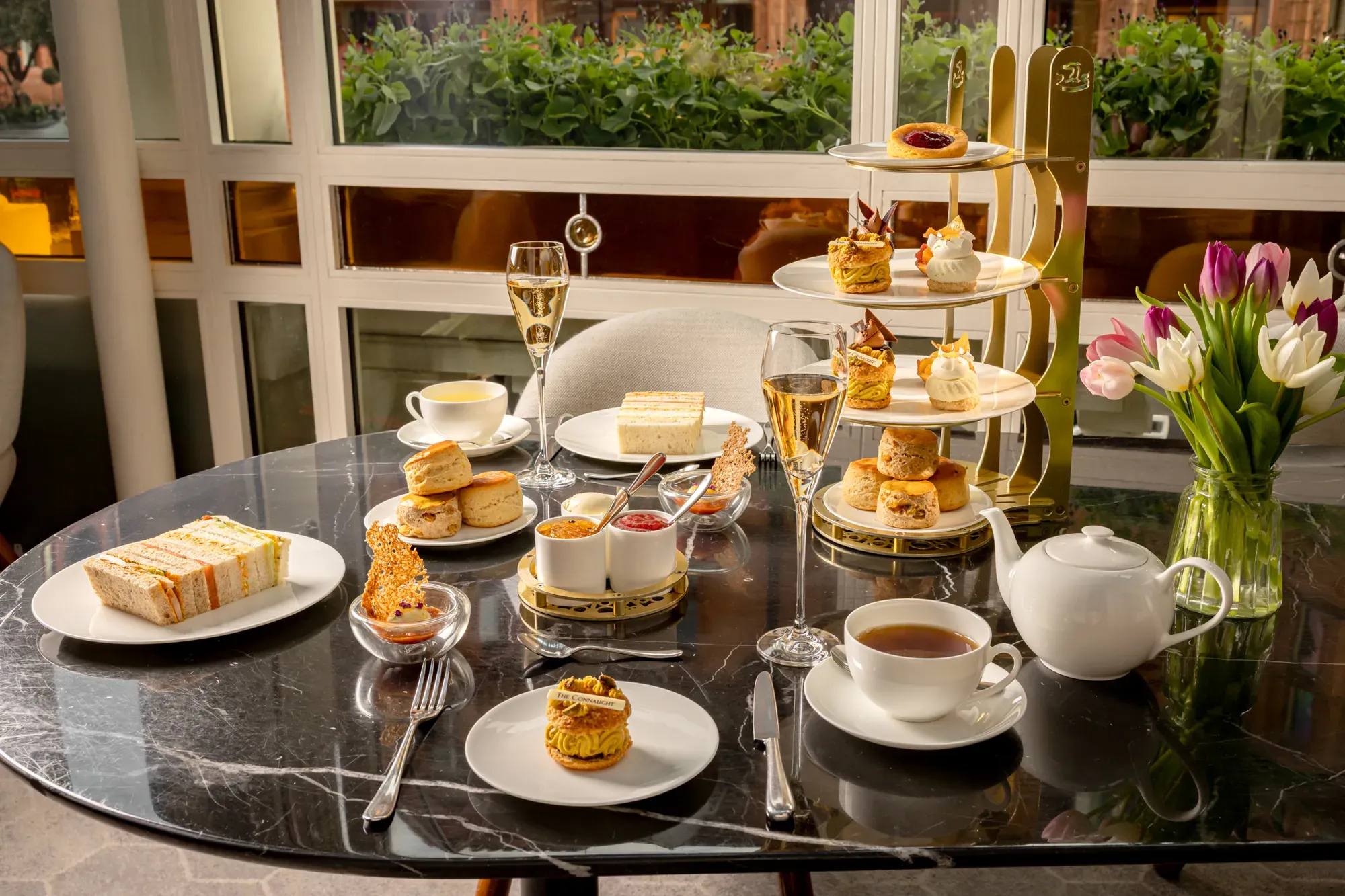 Afternoon tea spread with sandwiches, scones, pastries, tea, and champagne on a marble table beside tulips.