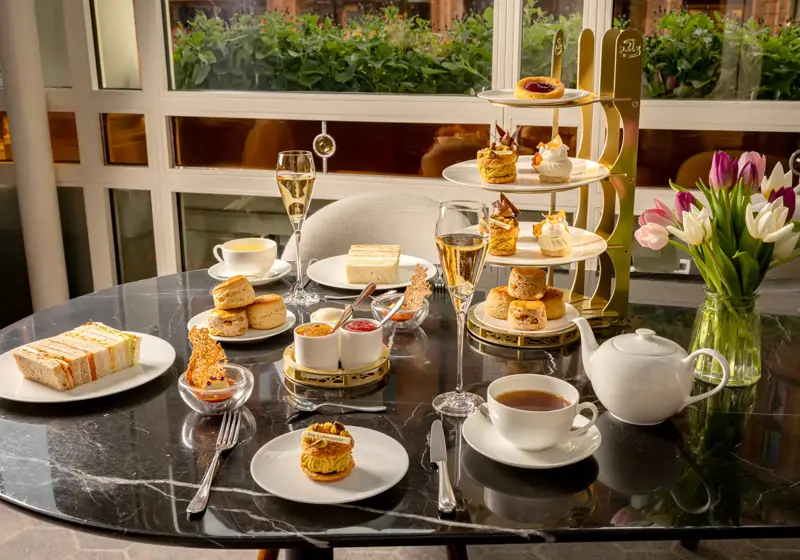 Afternoon tea spread with sandwiches, scones, pastries, tea, and champagne on a marble table beside tulips.