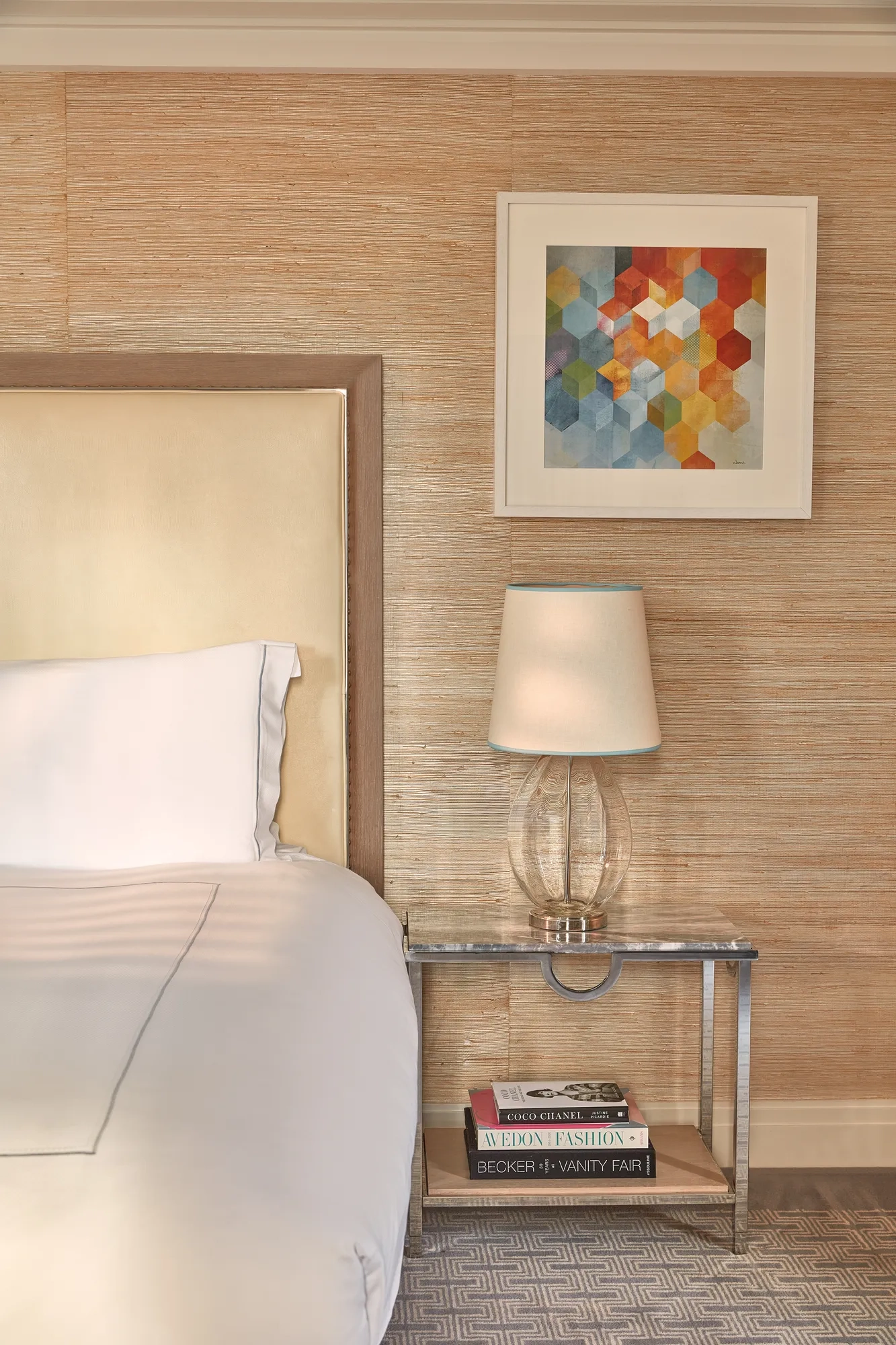 Close-up of bedside table with glass lamp, books, and abstract geometric artwork above cream upholstered headboard.