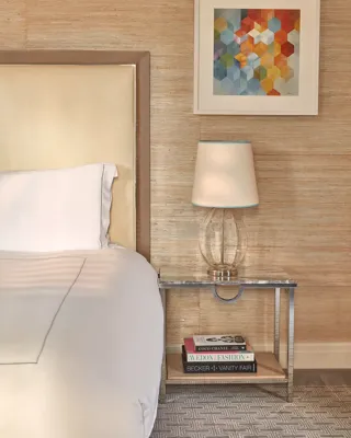 Close-up of bedside table with glass lamp, books, and abstract geometric artwork above cream upholstered headboard.