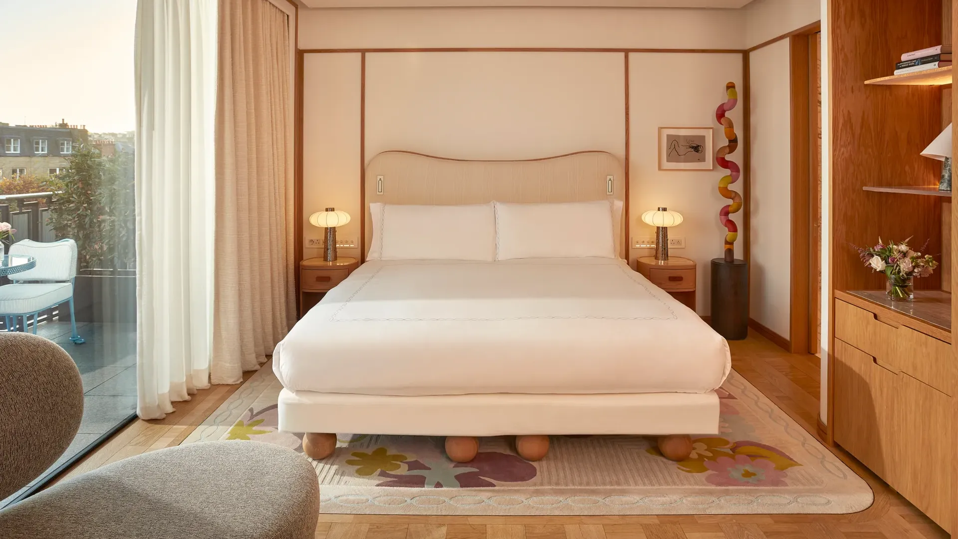 Bright bedroom with a king-size bed, wooden accents, and floor-to-ceiling windows opening onto a private terrace with outdoor seating.