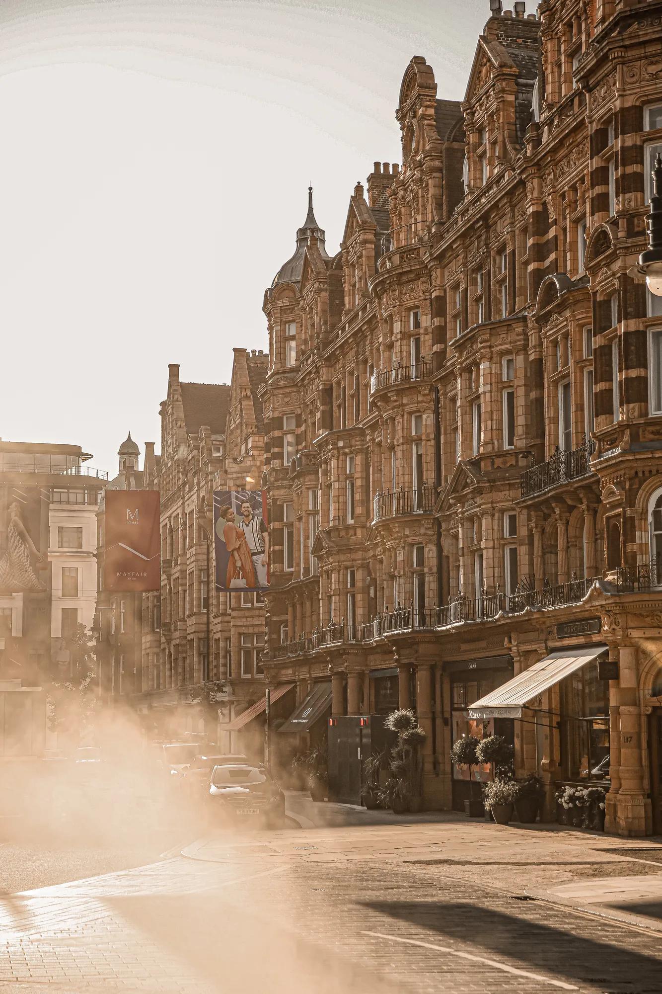 Sunlit historic street with ornate stone buildings, banners, and light mist rising from road, cars passing along warm-toned city scene.