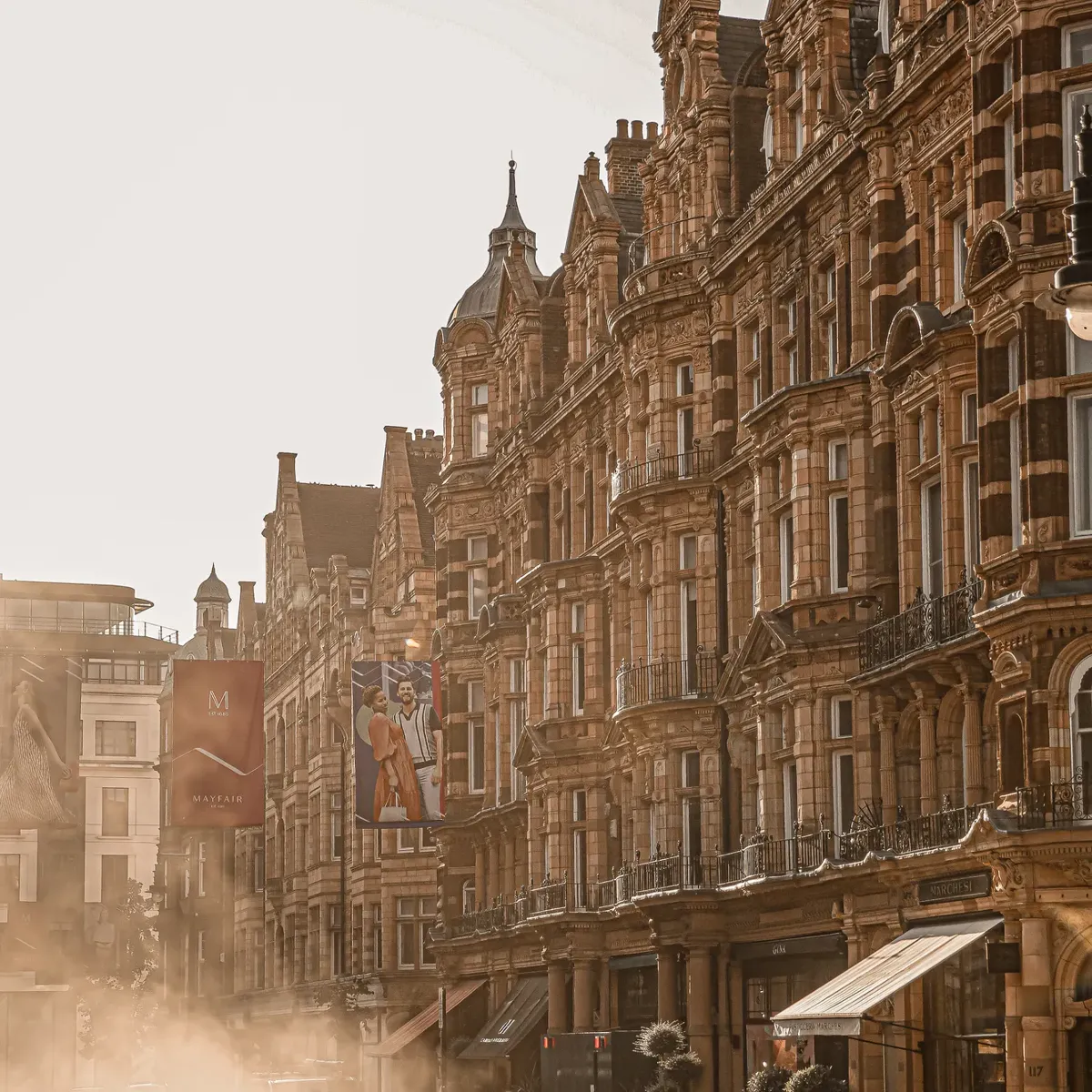 Sunlit historic street with ornate stone buildings, banners, and light mist rising from road, cars passing along warm-toned city scene.