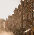 Sunlit historic street with ornate stone buildings, banners, and light mist rising from road, cars passing along warm-toned city scene.