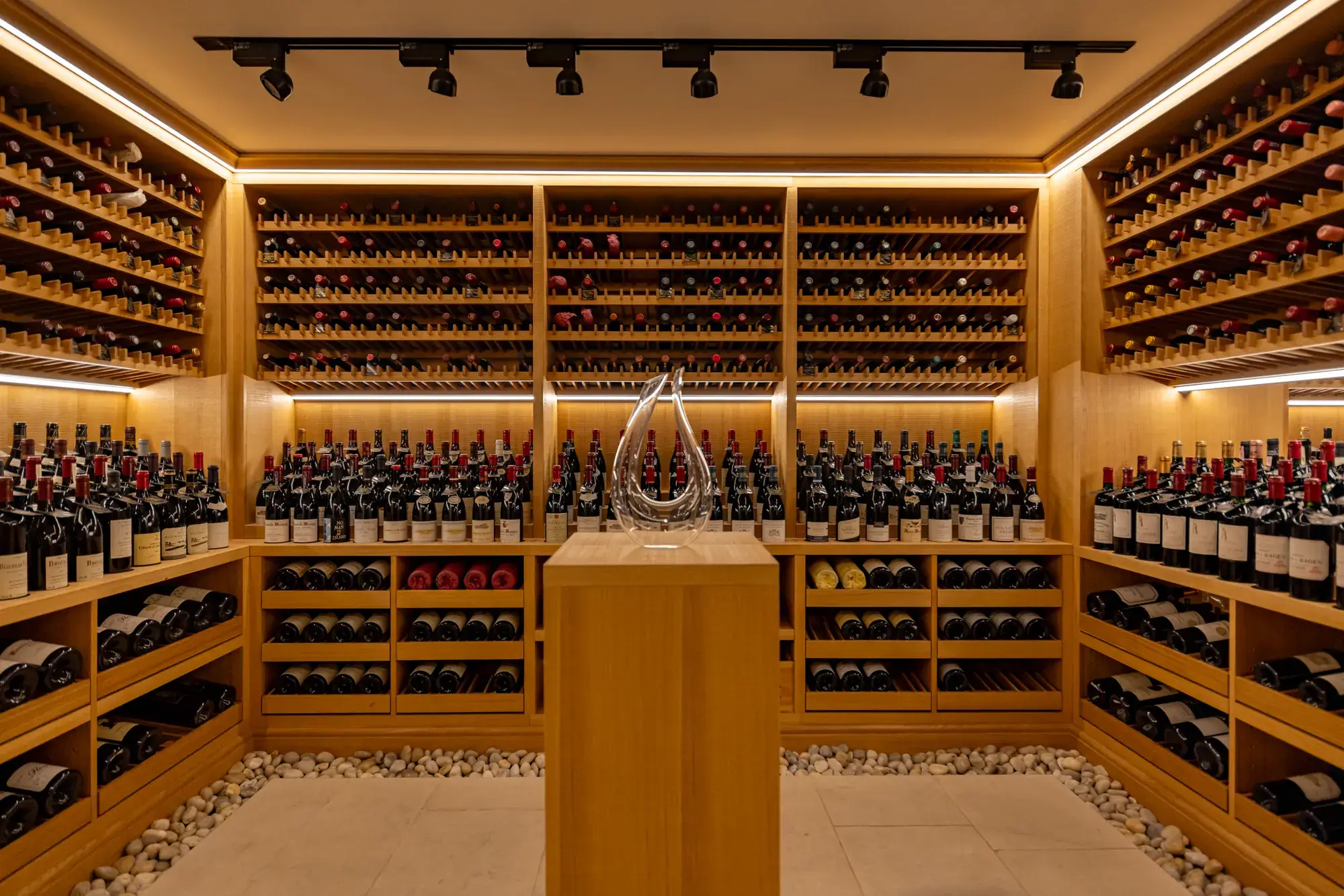 A softly lit wine cellar lined with wooden racks filled with red and white wine bottles, featuring a glass decanter displayed on a central pedestal surrounded by smooth white pebbles on the floor.