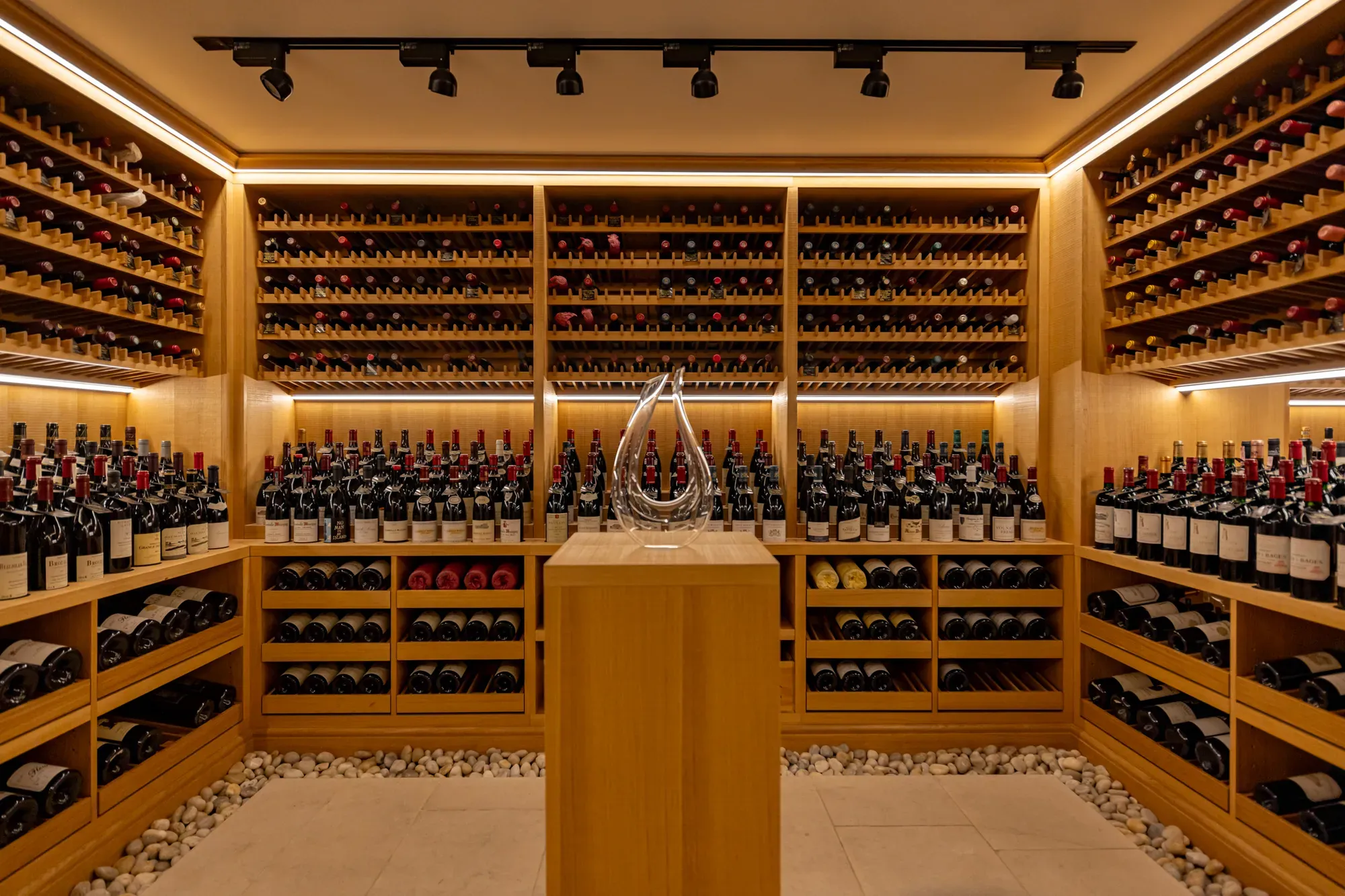 A softly lit wine cellar lined with wooden racks filled with red and white wine bottles, featuring a glass decanter displayed on a central pedestal surrounded by smooth white pebbles on the floor.