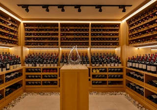 A softly lit wine cellar lined with wooden racks filled with red and white wine bottles, featuring a glass decanter displayed on a central pedestal surrounded by smooth white pebbles on the floor.