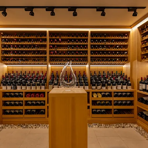 A softly lit wine cellar lined with wooden racks filled with red and white wine bottles, featuring a glass decanter displayed on a central pedestal surrounded by smooth white pebbles on the floor.