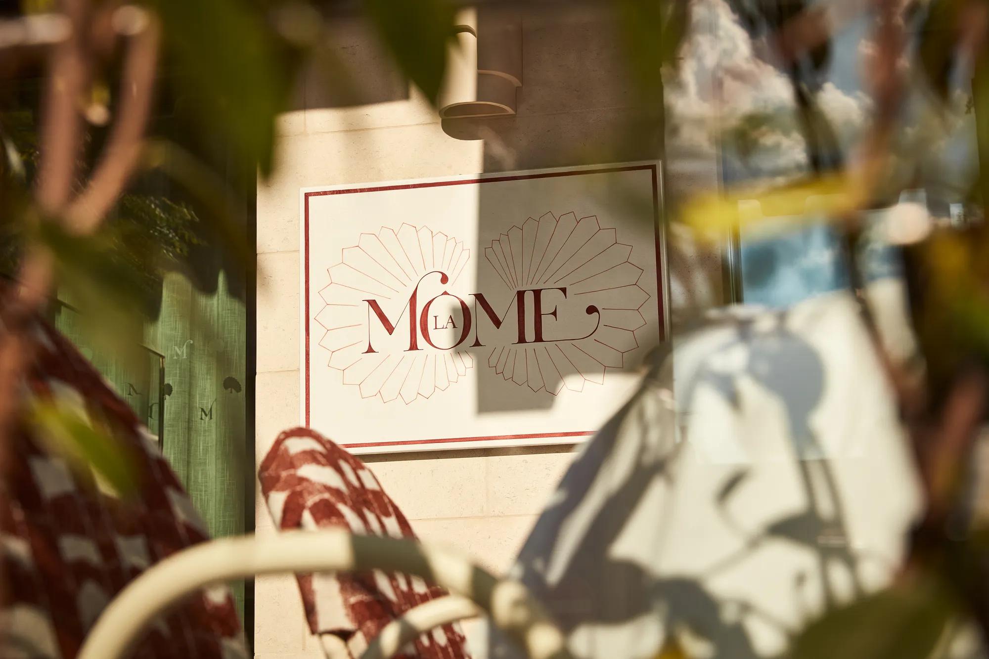 Close-up of La Môme restaurant sign framed by blurred foliage in foreground.
