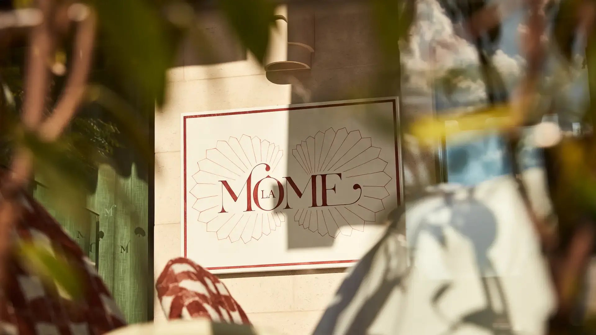 Close-up of La Môme restaurant sign framed by blurred foliage in foreground.