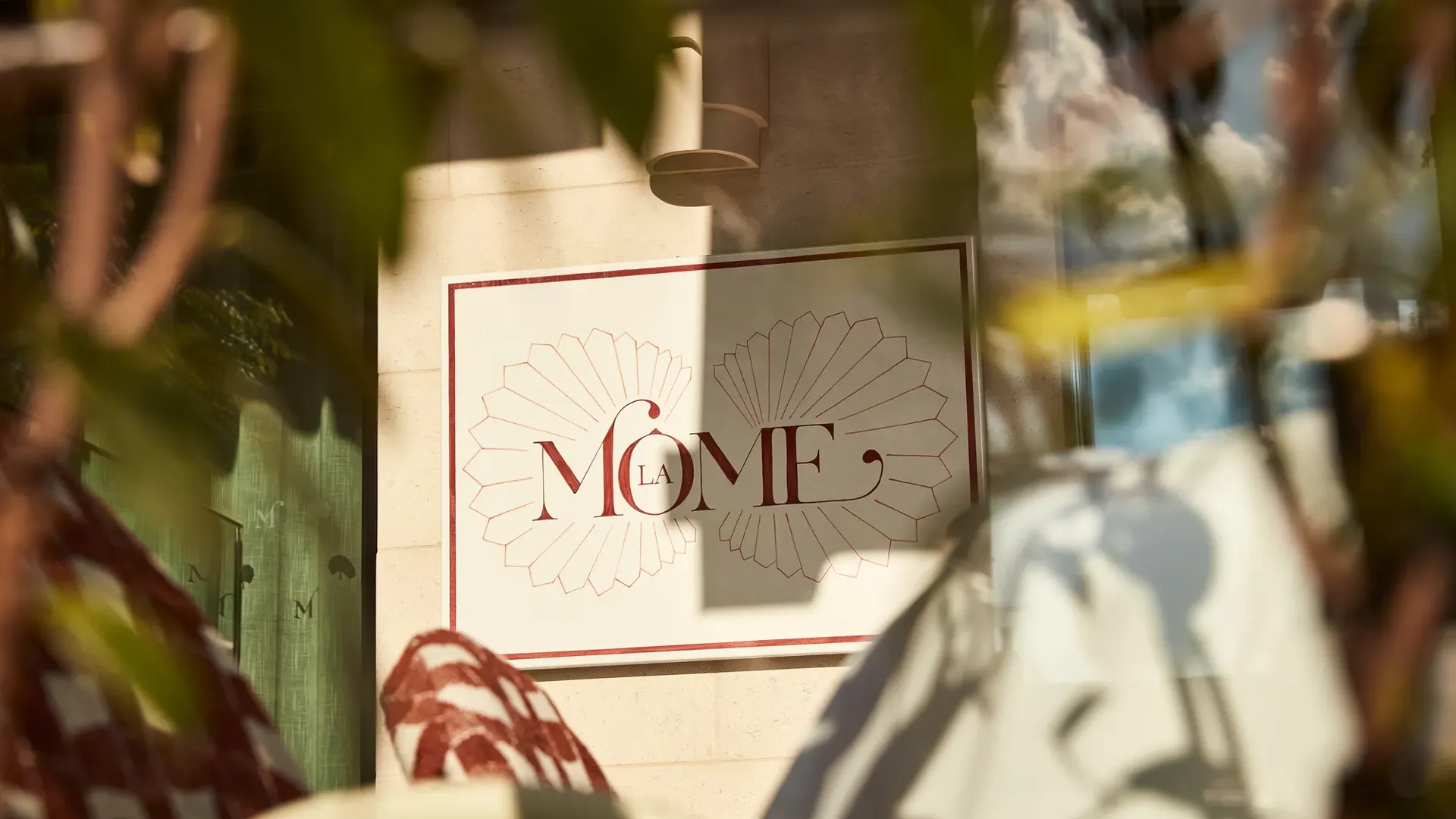 Close-up of La Môme restaurant sign framed by blurred foliage in foreground.