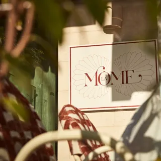 Close-up of La Môme restaurant sign framed by blurred foliage in foreground.