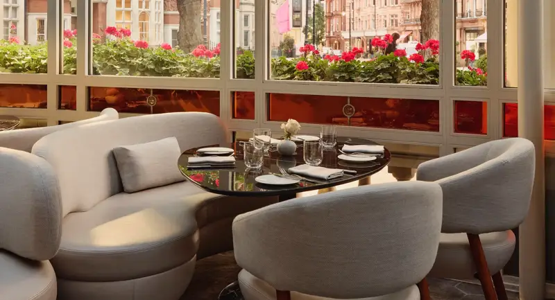 Bright dining alcove with curved white seating, round table, and large windows overlooking trees and flower boxes.