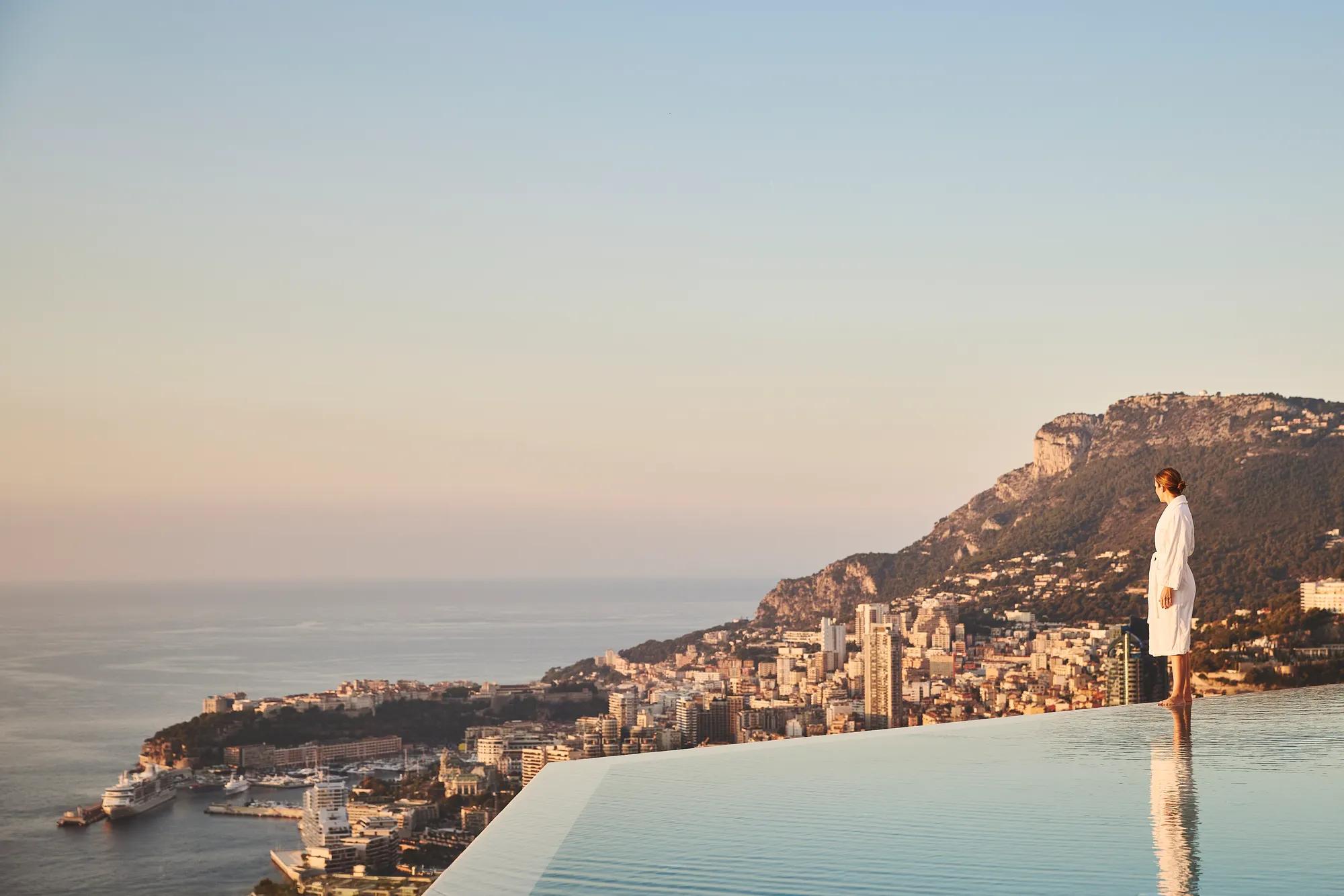 Person in white robe stands at infinity pool edge overlooking coastal city and sea, with mountains in warm evening light.