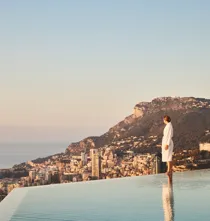 Person in white robe stands at infinity pool edge overlooking coastal city and sea, with mountains in warm evening light.