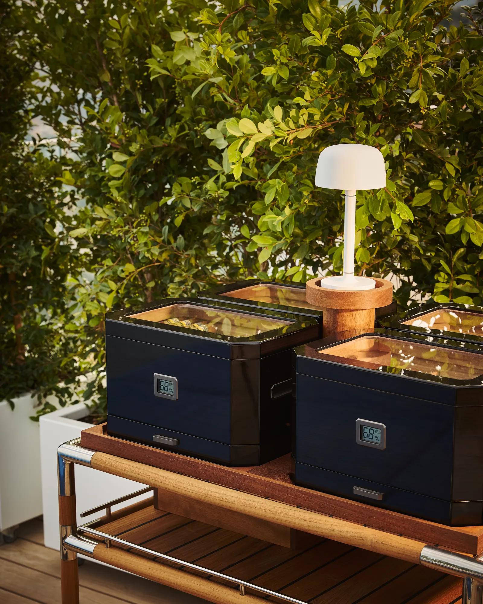 Outdoor buffet station with black food warmers on a wooden trolley, placed beside leafy green plants.