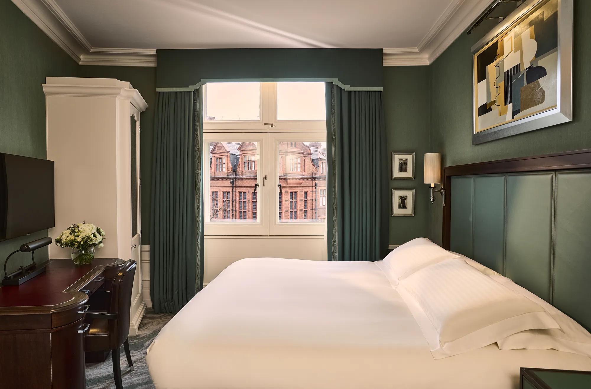 Elegant guest room with king bed, green walls, desk, wardrobe, and large window overlooking historic buildings.
