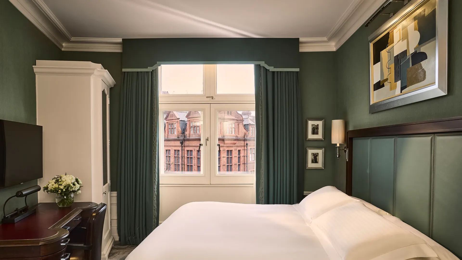 Elegant guest room with king bed, green walls, desk, wardrobe, and large window overlooking historic buildings.