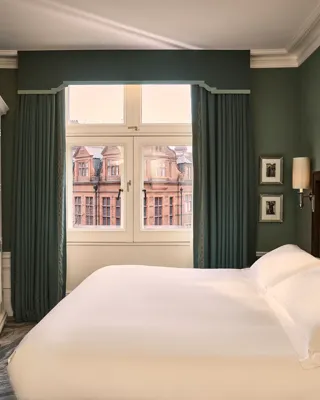 Elegant guest room with king bed, green walls, desk, wardrobe, and large window overlooking historic buildings.