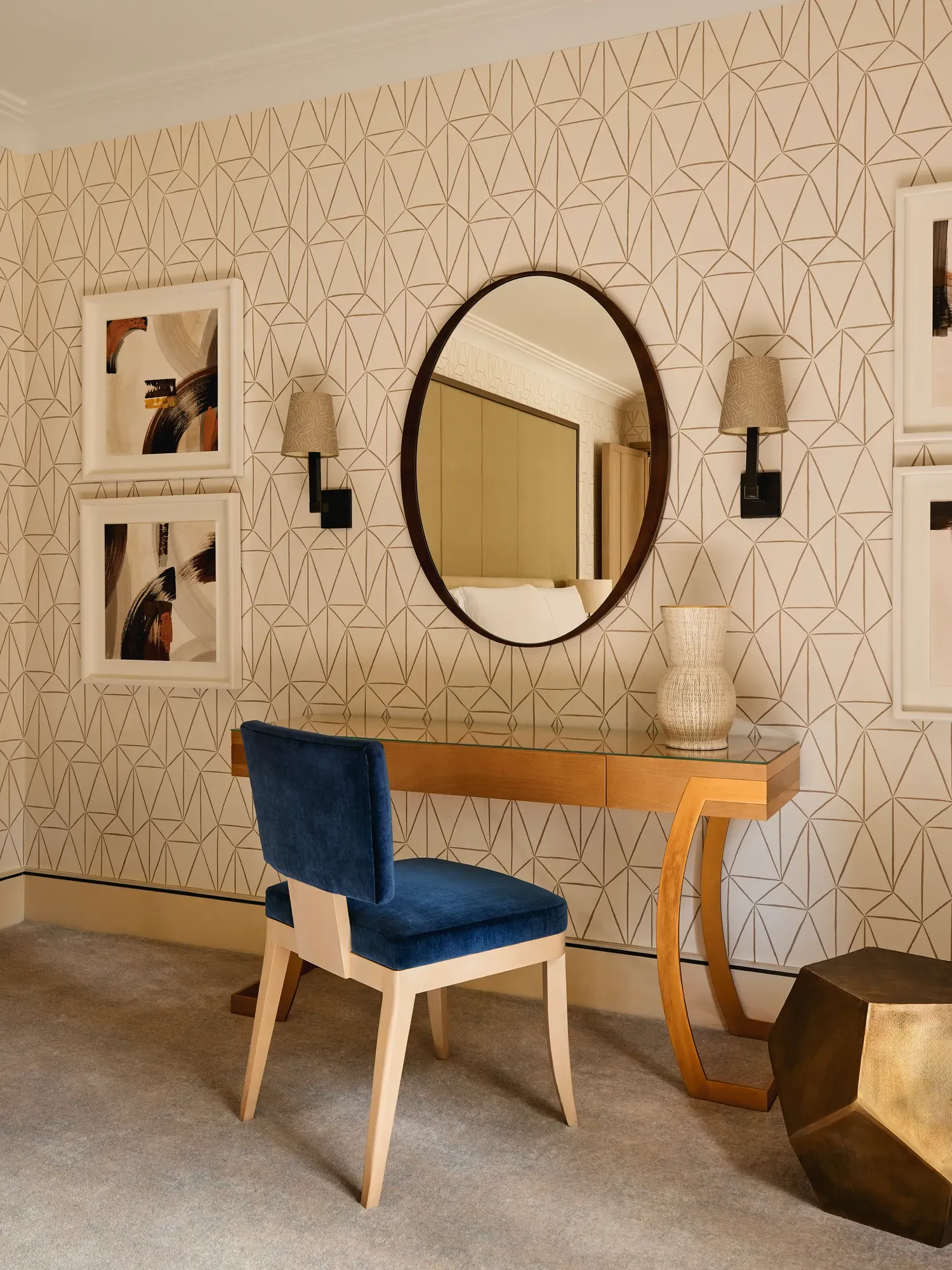 Elegant vanity area with blue chair, round mirror, patterned wallpaper, and framed artwork.
