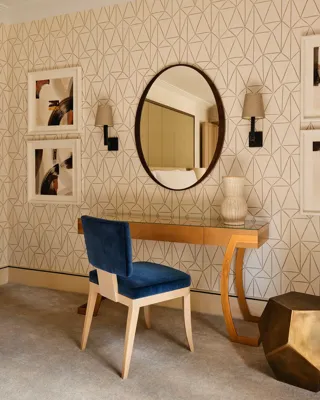 Elegant vanity area with blue chair, round mirror, patterned wallpaper, and framed artwork.