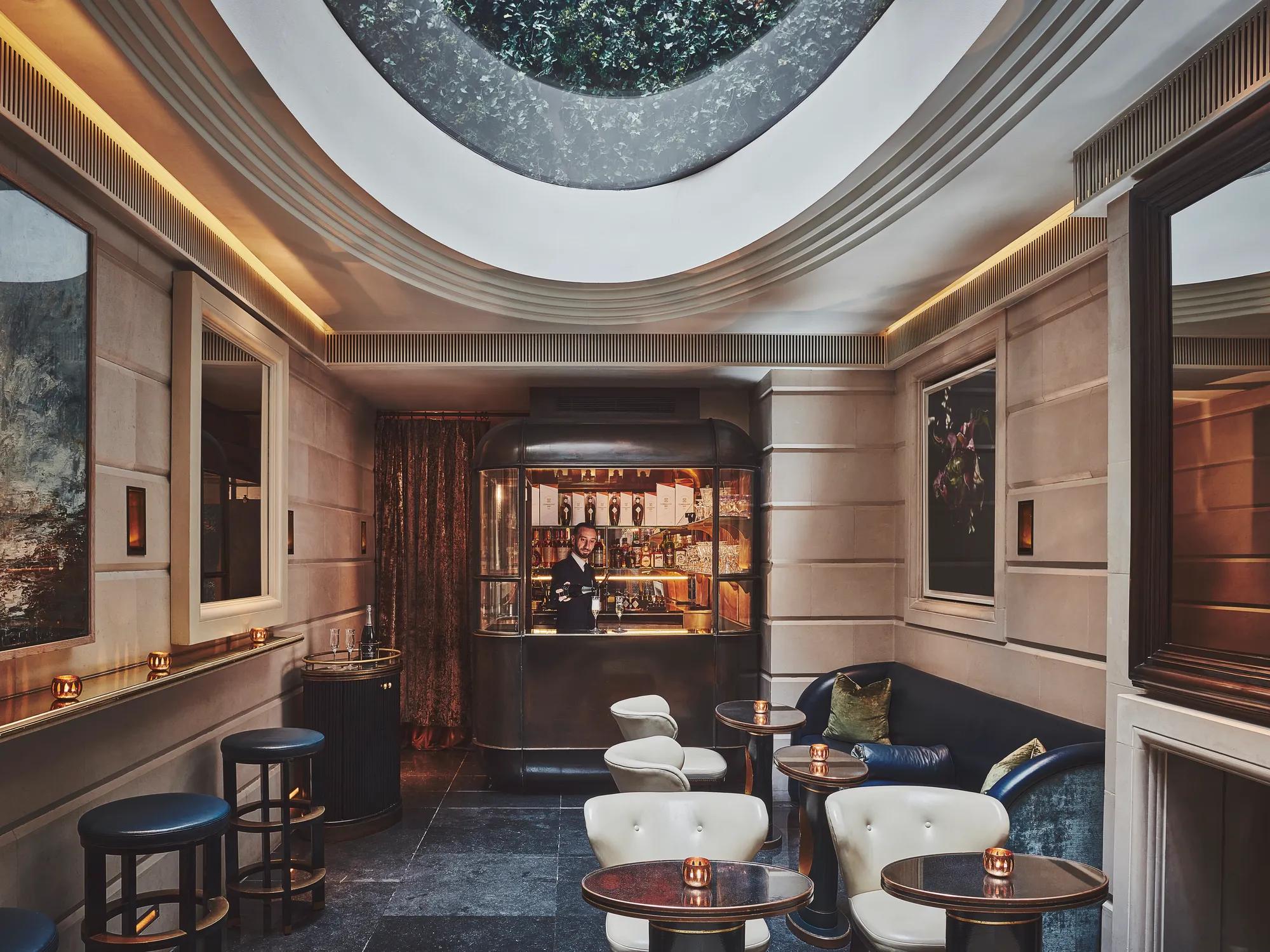 Intimate bar lounge with round tables, leather seating, and a skylight above, facing a small illuminated bar counter.