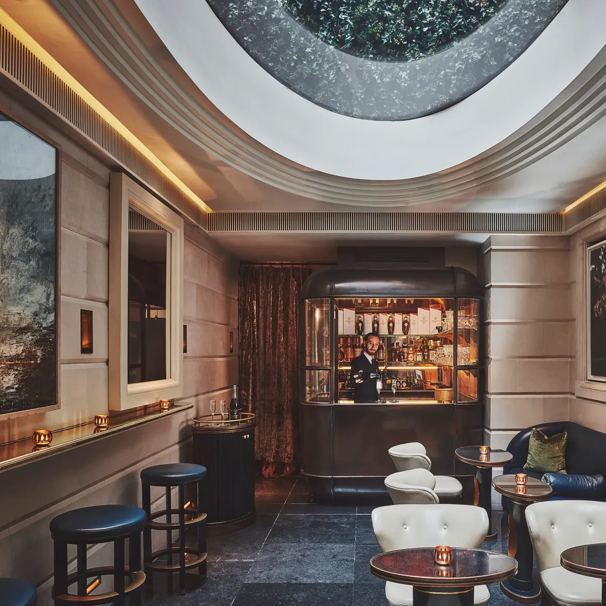 Intimate bar lounge with round tables, leather seating, and a skylight above, facing a small illuminated bar counter.